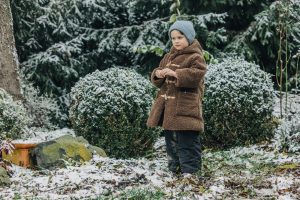 Little kid in hat and coat in winter snowy garden